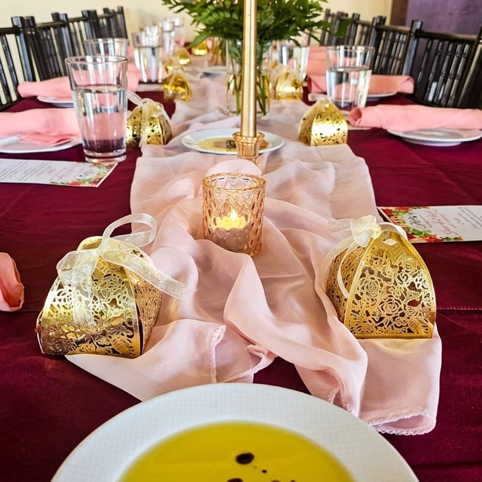 Festive restaurant table featuring burgundy linens, pink napkins, gold lanterns, candles, water glasses, and a bowl of soup.