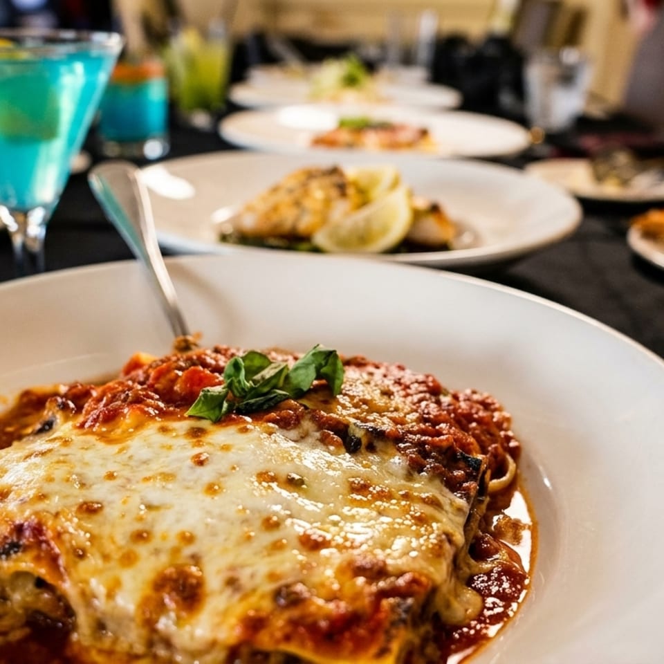 Cheesy lasagna garnished with basil in the foreground, surrounded by plated dishes and a bright blue cocktail at a restaurant table.