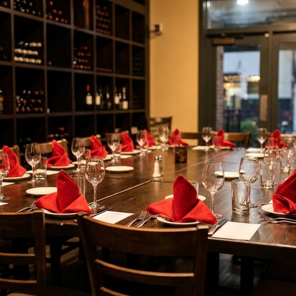 Elegant restaurant dining room with a long wooden table set with red folded napkins, wine glasses, and bottles displayed on shelves.