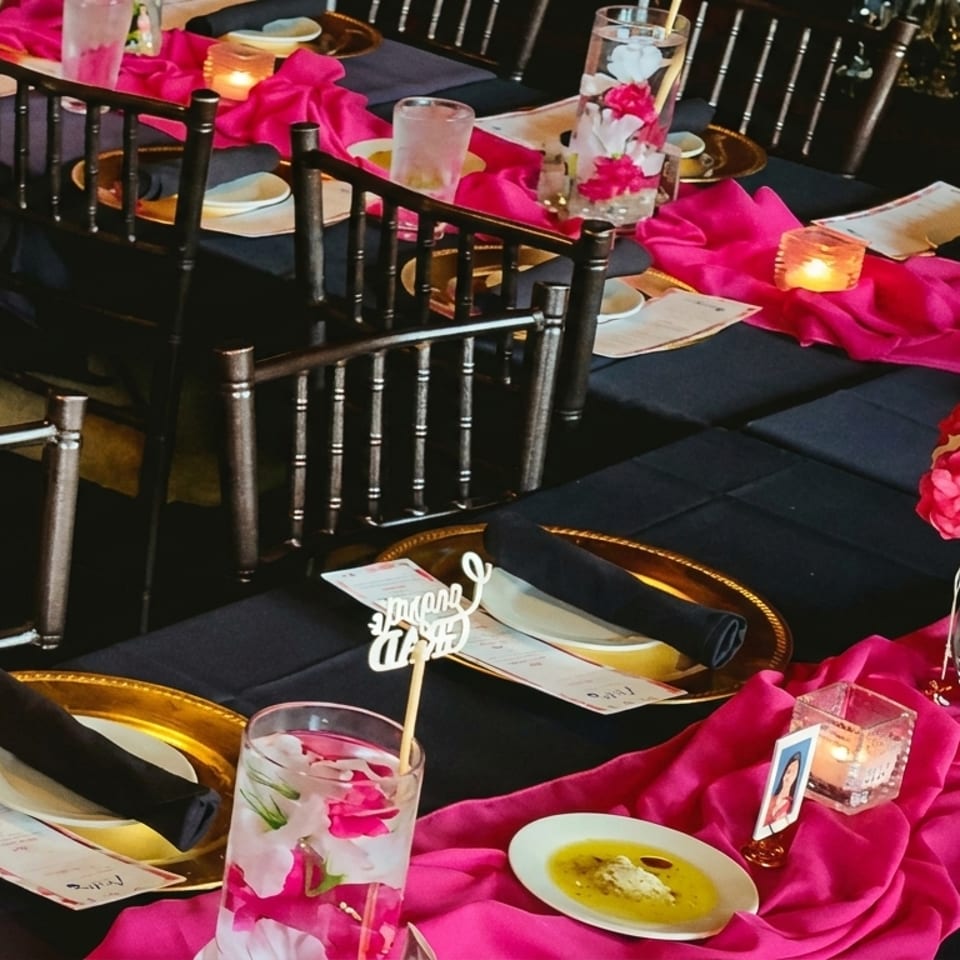 Elegantly set banquet tables feature black linens, hot pink runners, roses, candles, gold chargers, and place settings.